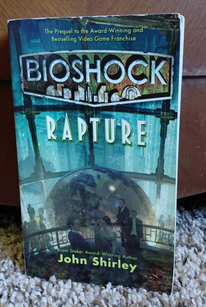 Image is a photo of a book cover for Bioshock Rapture by John Shirley. It shows a huge party room with the outside of the windows being under water. There's a big globe in this room with an upper balcony area. There are several people on the cover in fancy attire. 