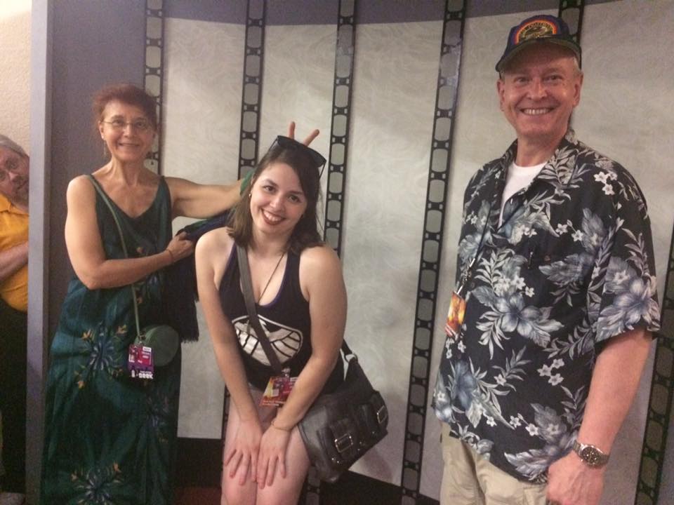 [Image is a photo of 3 people. The person on the left is doing bunny ears to the person in the middle. The person on the right is smiling]