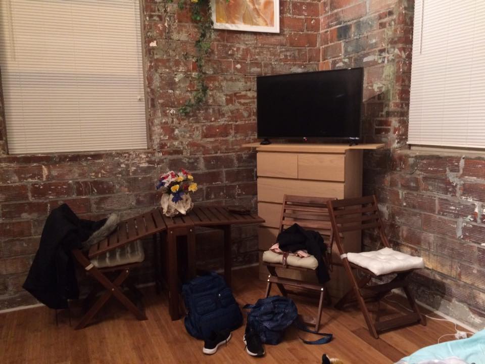 [Image show s a red brick wall that looks aged and broken down. There's a dresser where a small TV sits. There are several chairs and a small table with flowers on it]