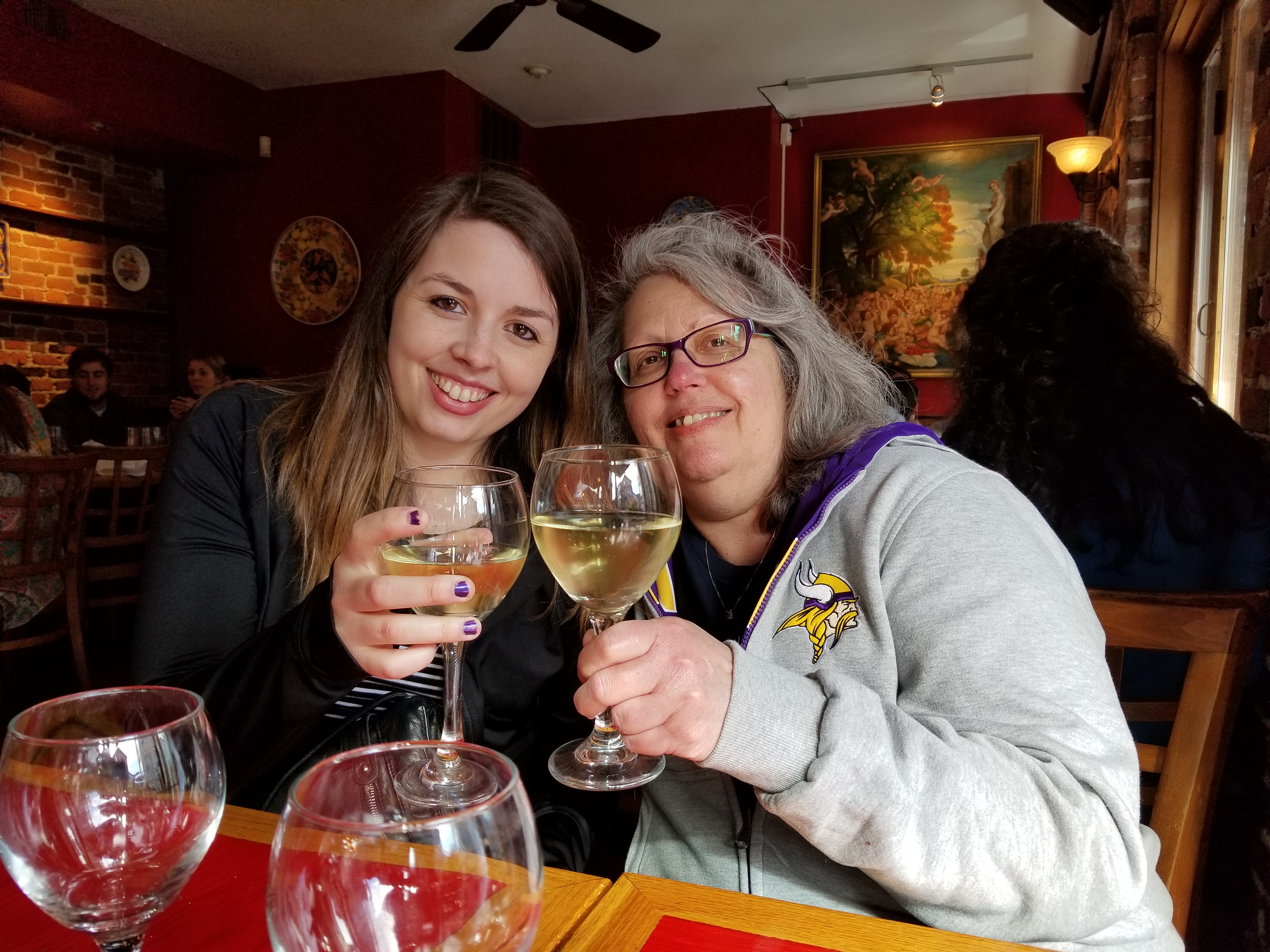 Image is a photo of me and my mom from our trip to Boston in 2018. We are having dinner and holding up our wine glasses. We are smiling at the camera. 