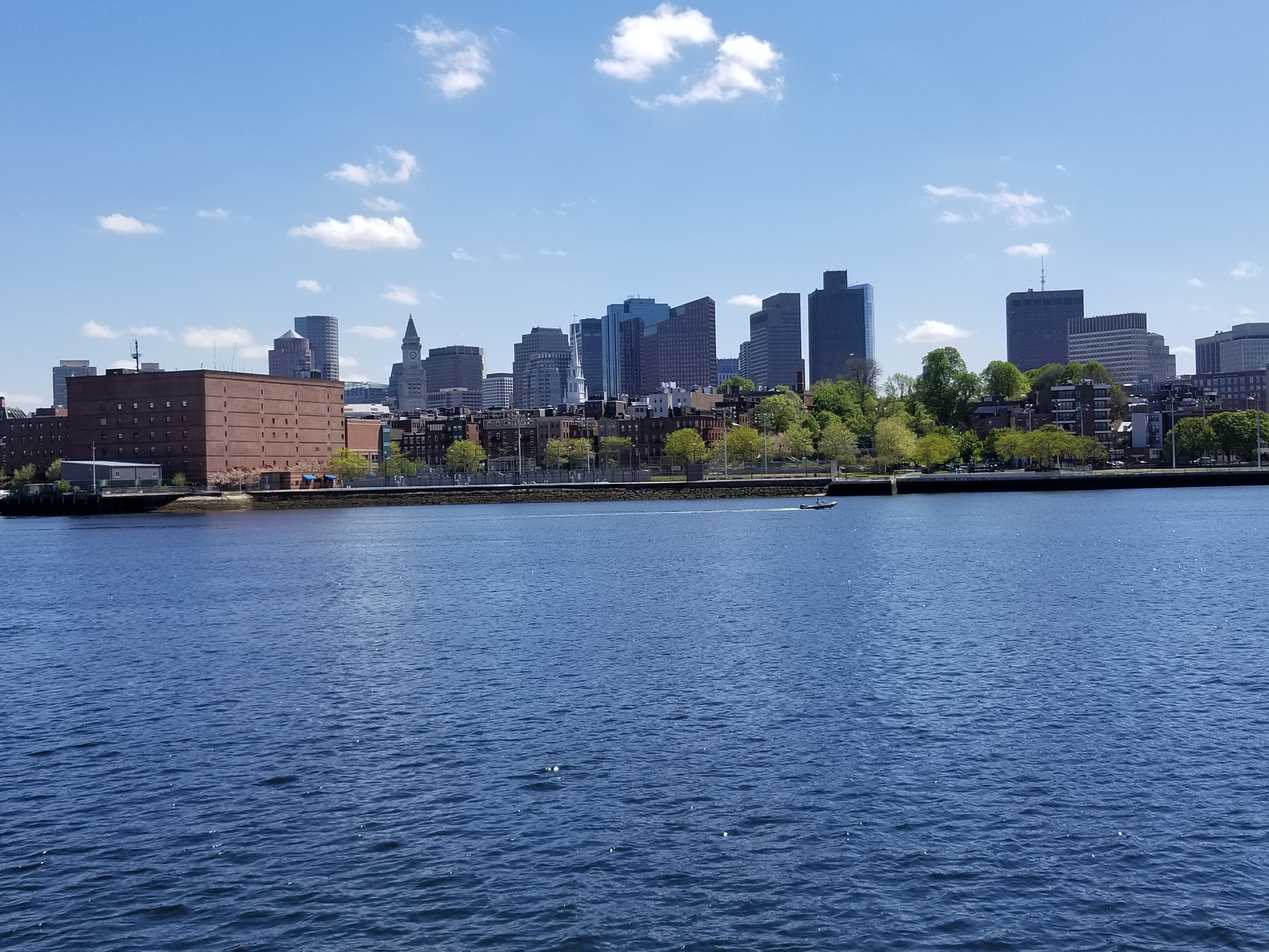 [Image is of the Boston skyline. The water is blue, the trees are really green. There's a boat in the water passing by]