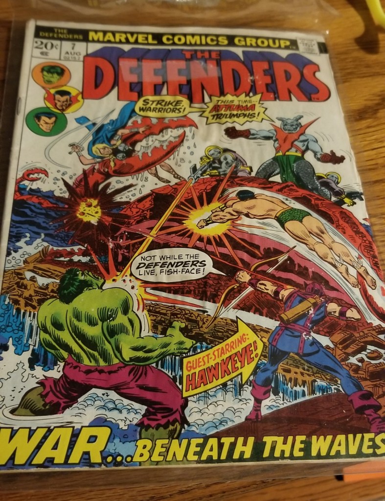Image is a photo of a comic called The Defenders. On it is the Hulk, Hawkeye with his bright blue and purple outfit. It show other characters fighting together. 