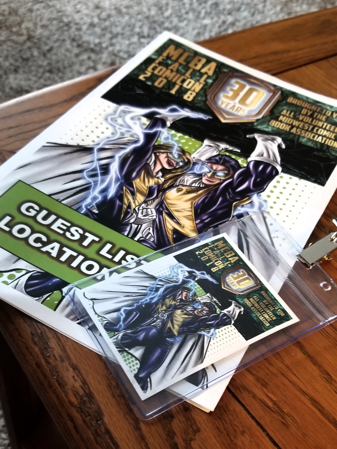 [Image is a picture of a convention book and a badge. It says MCBA Fall Con 2018 - 30 years. It shows two superheroes with lightning coming out of their eyes holding up part of the sign]