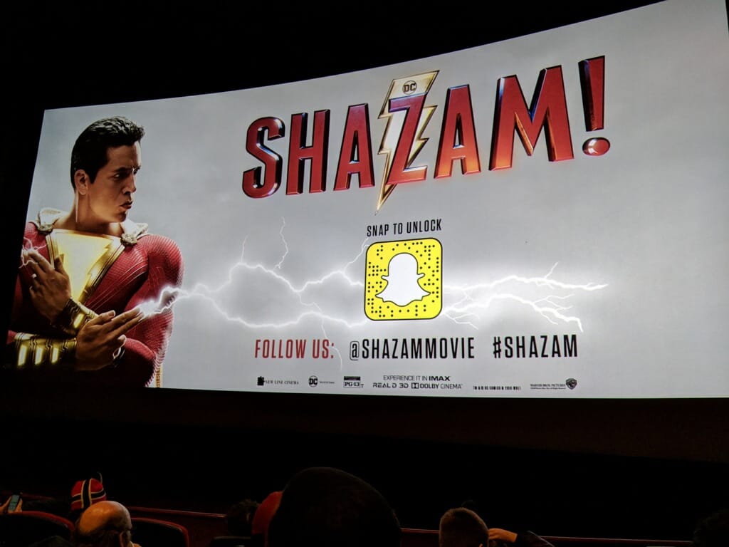 Image is a move theater with a man to the left. He's pointing his finger and lightning comes out of it. It shows the movie logo Shazam with a lightning bolt behind the Z.