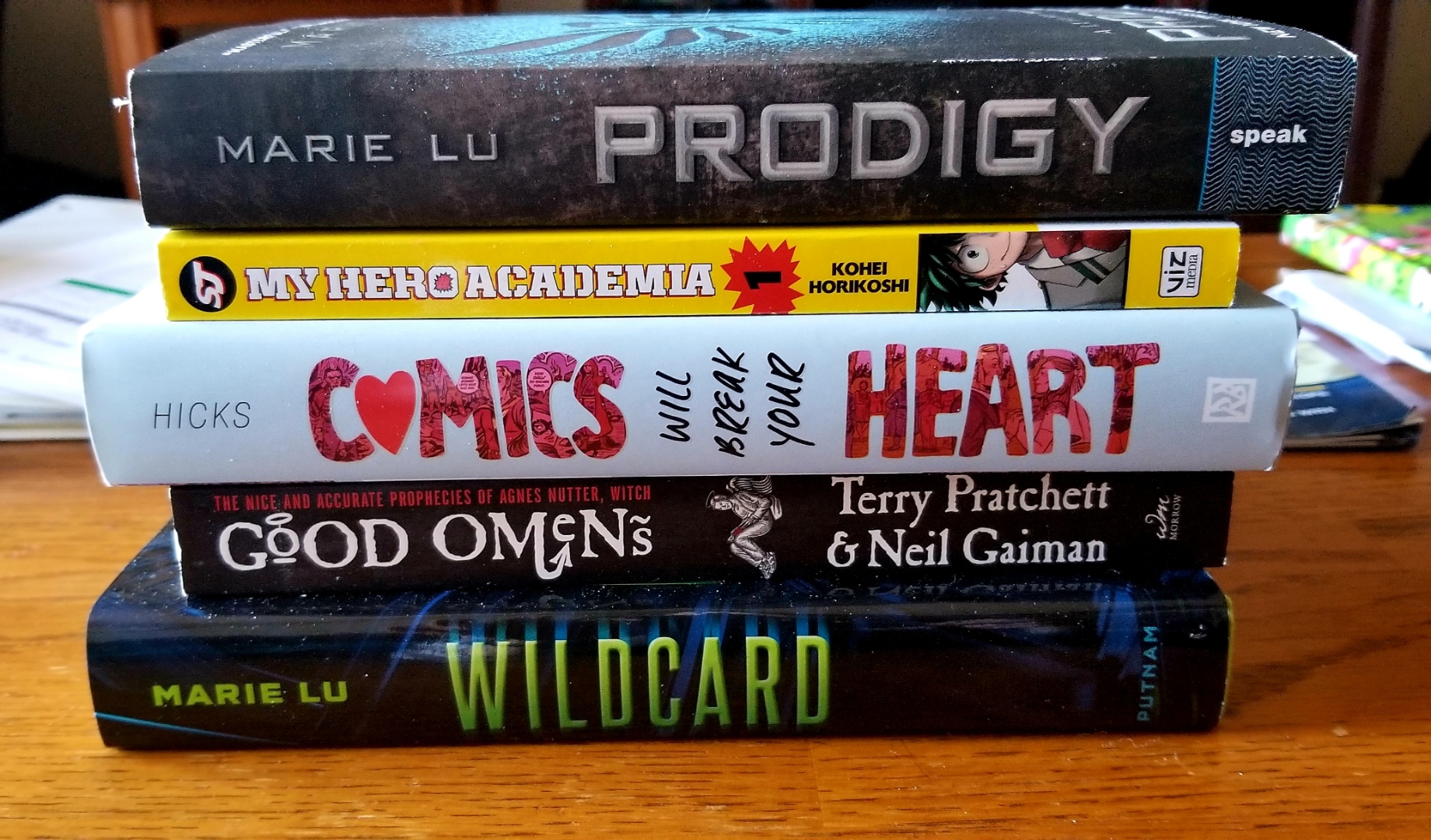 Image is a stack of five books. From top to bottom, they are: Marie Lu Prodigy, My Hero Academia #1, Comics Will Break Your Heart, Good Omens, and Wildcard. 