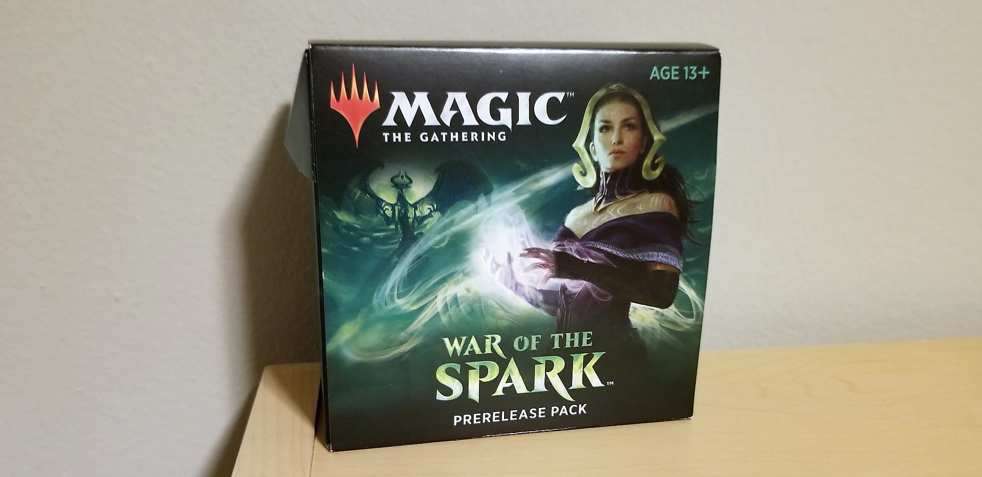 Image is a photo of a Magic: The Gathering pre-release kit. It shows a woman holding purple magic in her hands. At the bottom it says "Wr of the Spark prerelease pack"