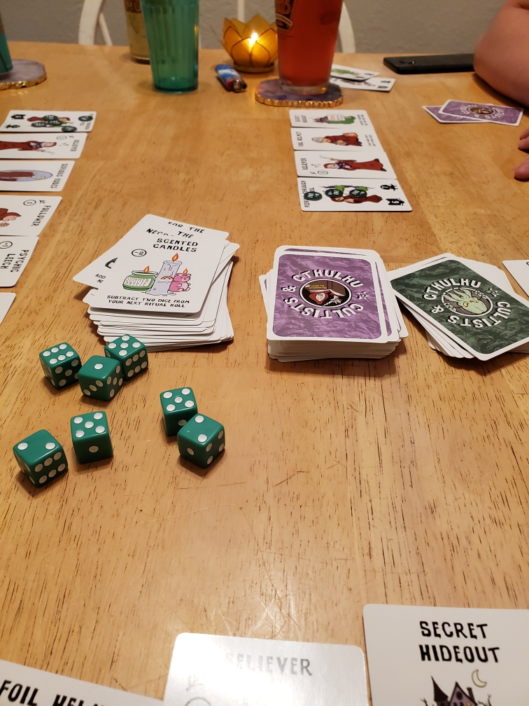 Image is a photo of cards on a wood table. There is a left discard pile. The deck which says Cultists & Cthulhu and a dark green deck next to it. There's also several green dice to roll nearby. 