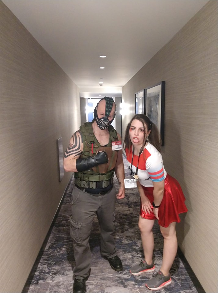 Image is a photo of a Bane cosplayer and a Harley Quinn cosplayer in a hallway of a hotel. Bane has his full mask on from The Dark Knight Rises and Harley is sticking her tongue out at the camera. 