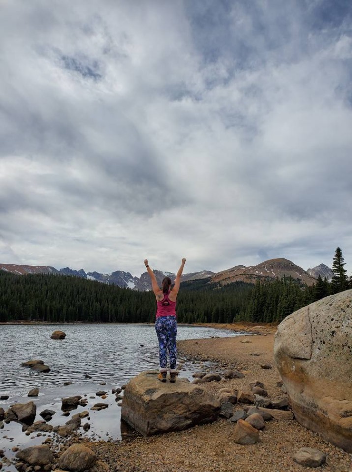 Image is a photo of me after completing a hike. This photo shows my back with both my arms raised above my head in triumph. There's a forest and stream ahead of me with the mountains further in the background. 