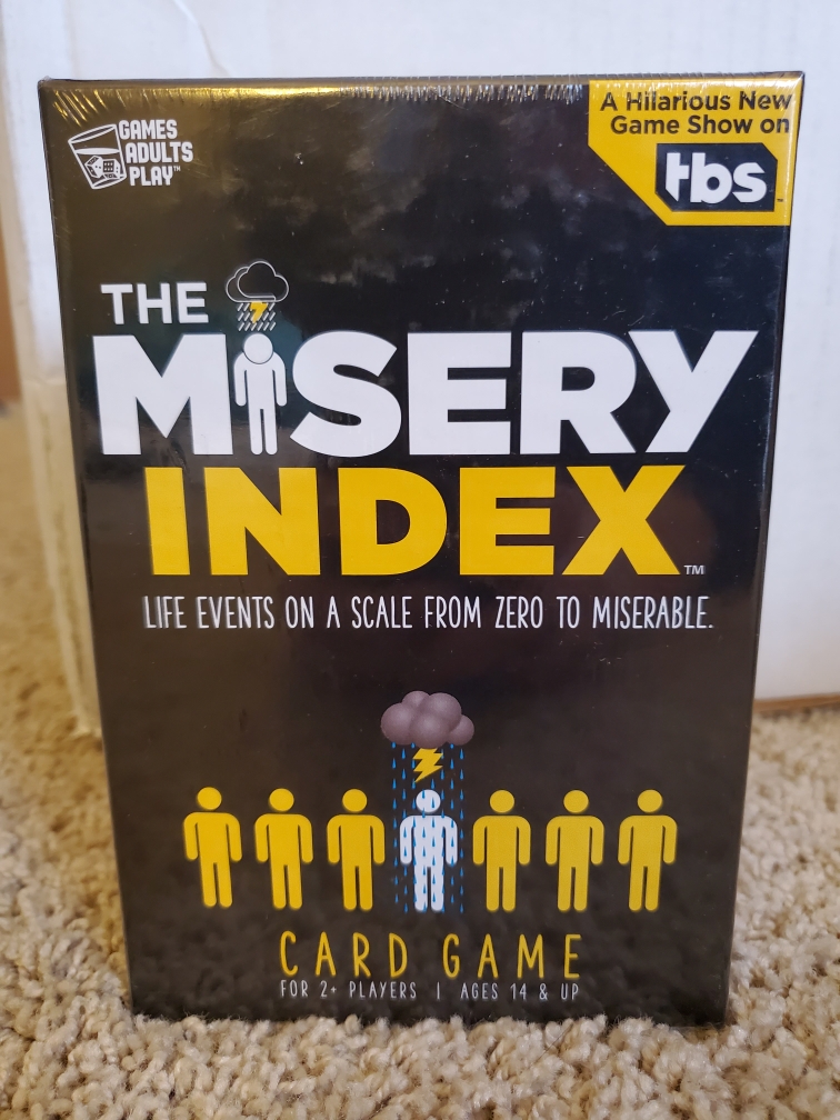 Image is of the board game The Misery Index. It shows cartoon people. The I in Misery Index is a person who looks sad. On the bottom are seven cartoon people. All of them are yellow except on in the middle is white. There's a rain storm over the white cartoon person. 