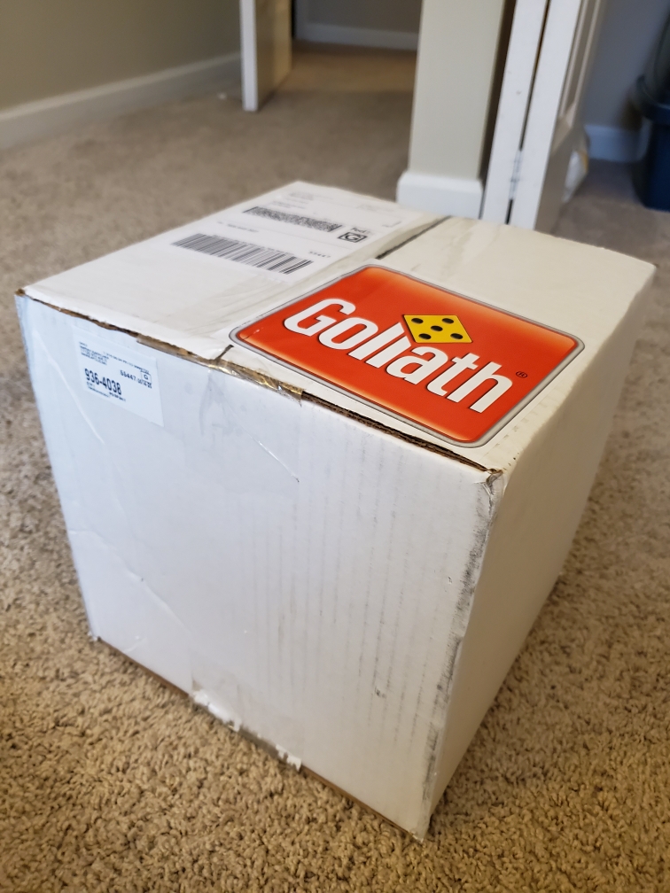 Image is a photo of a white box that is wrapped and was shipped to me. On the white box is a logo for Goliath. On the L and the I is a yellow and black d6 die. 