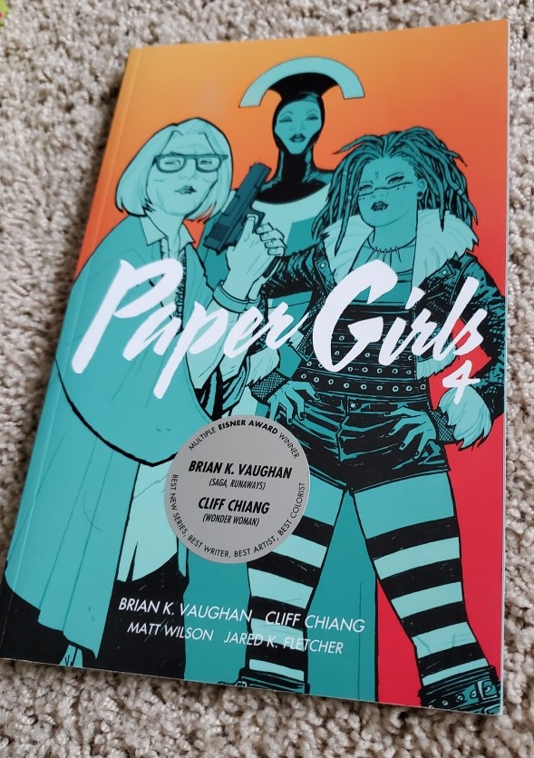 Image is a photo of the comic book series, Paper Girls. This is volume #4. It shows three characters - one is older and has a bob. She's holding a hand gun. The character in the back is wearing an eccentric head piece that's a half circle. The woman on the right has what appears to be dreads and tribal face paint on. 