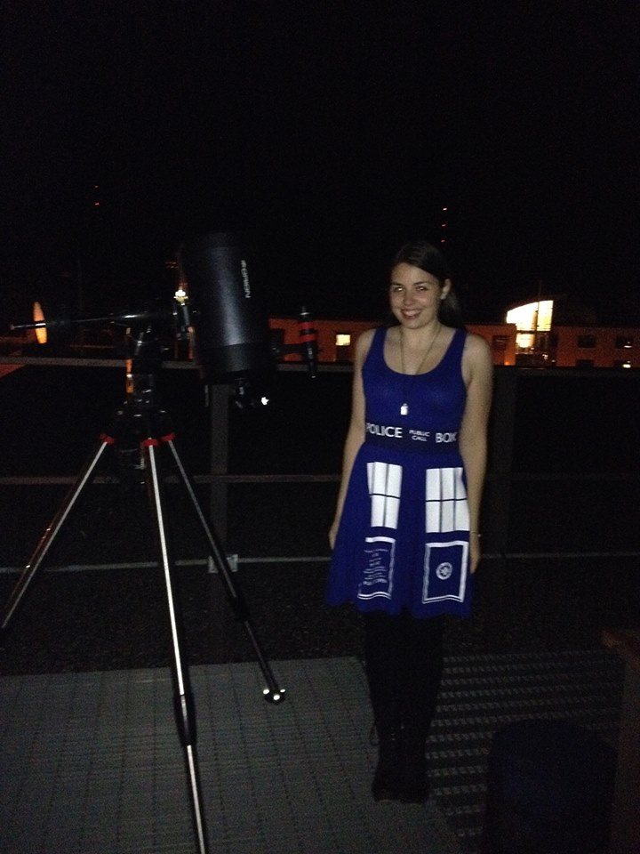 Image is a photo of me taken at night. I'm wearing a Tardis dress (a blue telephone booth from Doctor Who). I'm smiling and next to me is a telescope. 