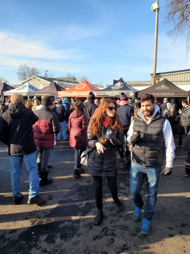 Image is a photo of several people standing in front of beer tents. On each tent is a different brewery. Everyone is wearing coats and hats trying to keep warm. The sky is blue. 