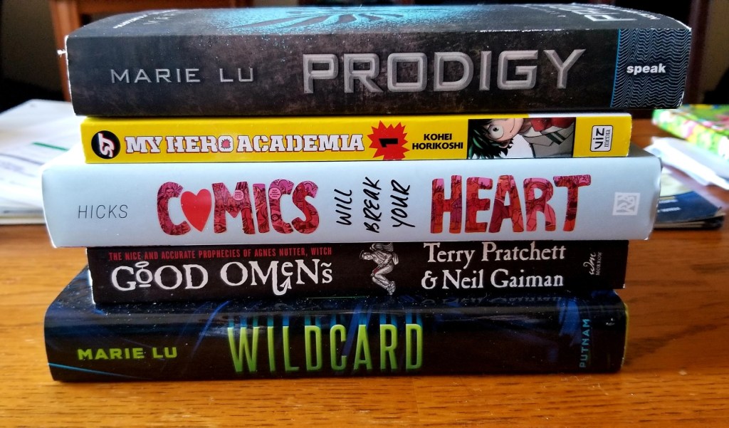 Image is a stack of five books. From top to bottom they are: Prodigy by Marie Lue, My Hero Academia 1 by Kohei Horikoshi, Comics will break your heart by Faith Erin Hicks, Good Omens by Terry Pratchett and Neil Geiman, and Wildcard by Marie Lu