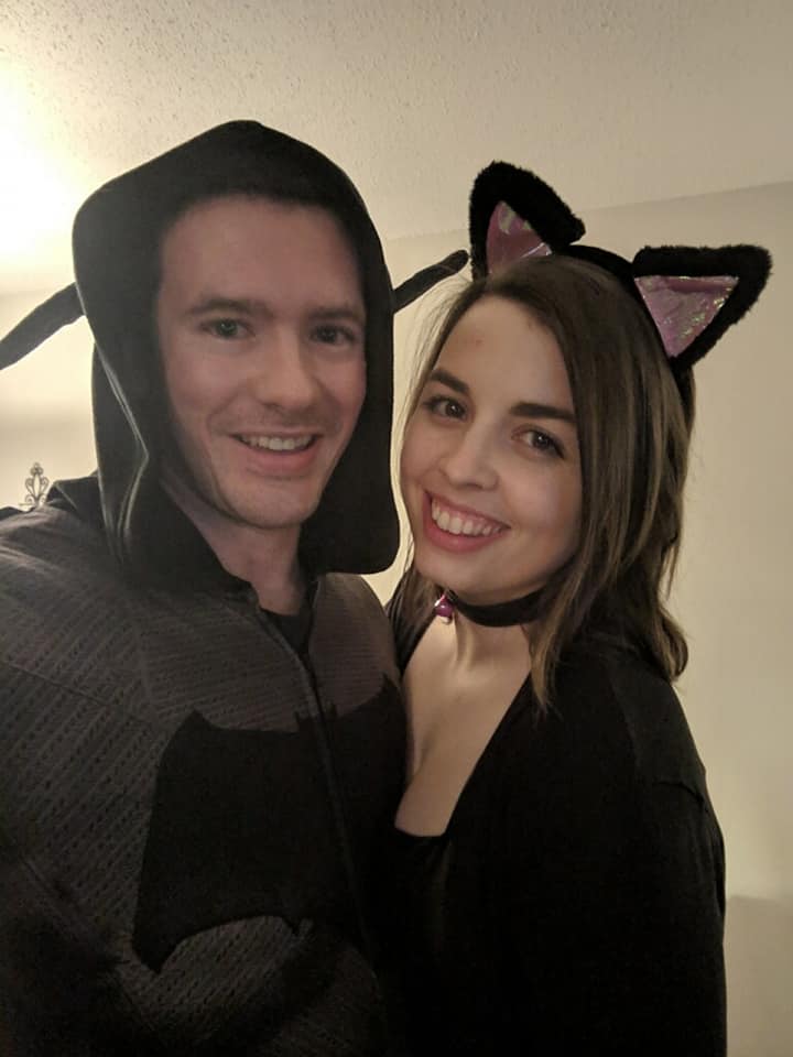 Image is a photo of me and my fiance. He is wearing a Batman oneside and I'm wearing cat ears. We are both smiling at the camera. 