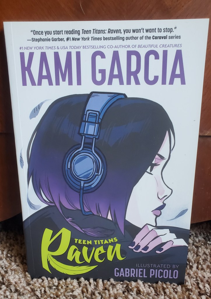 Image is a photo of a graphic novel book cover for Kami Garcia and Gabriel Picolo's graphic novel, Teen Titans Raven. It shows a woman's side portfolio as she's looking ahead. She has a purple bob with noise cancelling head phones on her head. 
