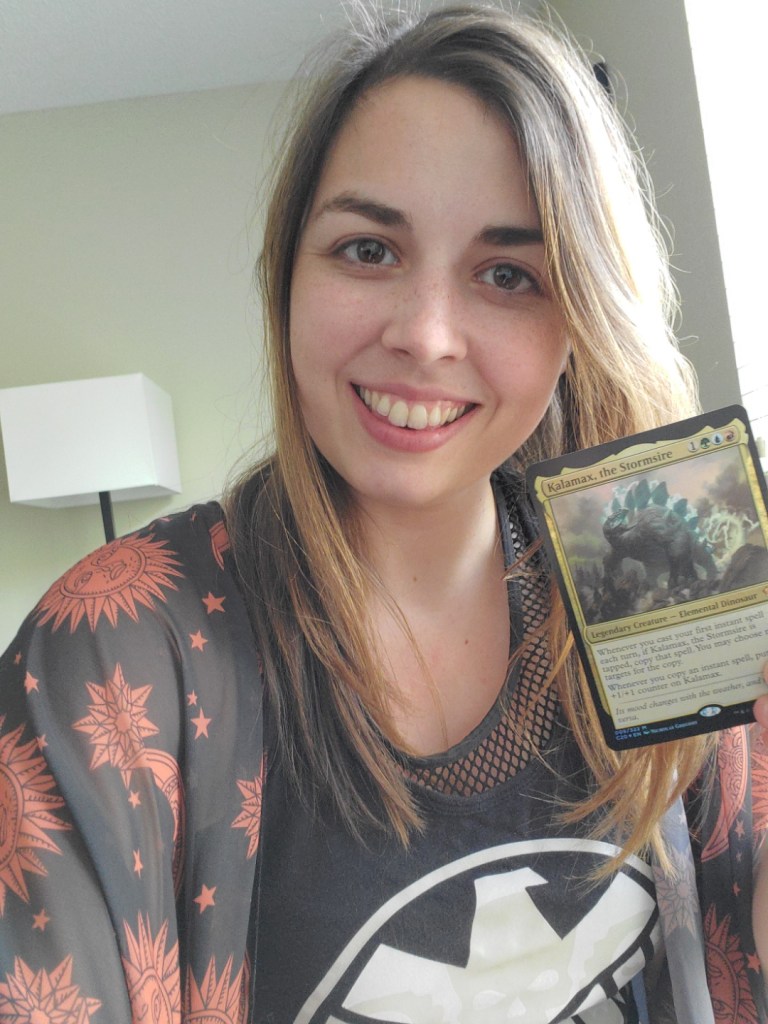 Image is a selfie of me holding a Magic card. I'm wearing a black and orange shawl with my Agents of Shield tank top. My hair is a blonde mixed with brown. The card is "Kalamax, the Stormshire". It's a gray and blue dinosaur with lightning coming from its spine. 