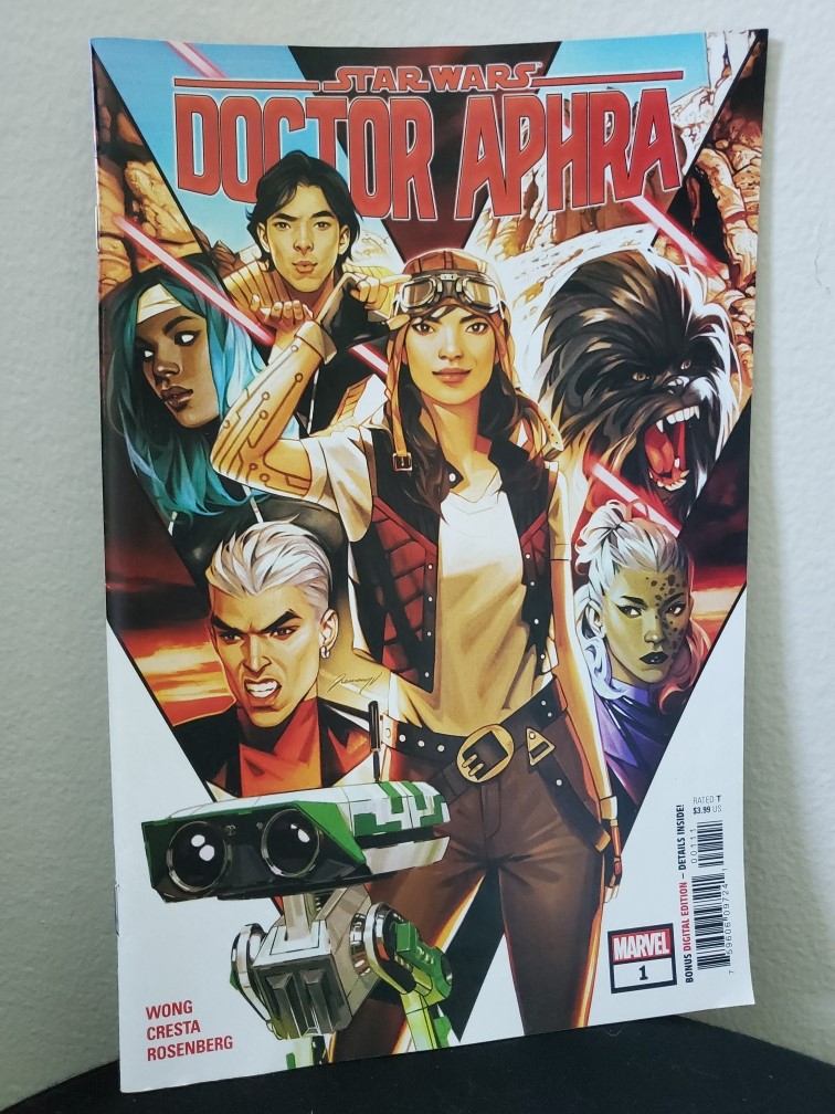 Image is a comic book cover for Doctor Aphra Issue #1. It shows a woman on the cover. She's wearing a white t-shirt, red vest, and a hat with what looks like ski goggles on her head. There are several other characters surrounding her. 
