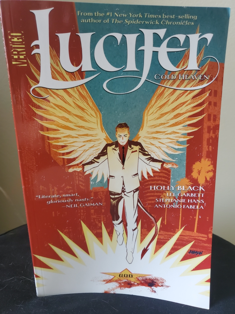 Image is a photo of the comic book cover for Lucifer Cold Heaven. It shows a man glowing and he is wearing a gray suit with a red shirt and black tie. He has white angel wings. It shows him landing on a Hollywood star that has God in the middle of the yellow star. There are blood stains on the star. 