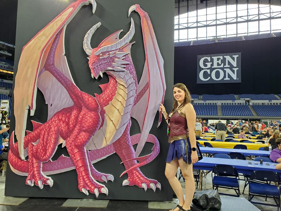 [This picture features a big cut out of Genieveve the dragon logo of Gen Con. I'm dressed as Wonder Woman and I'm pointing at the dragon]