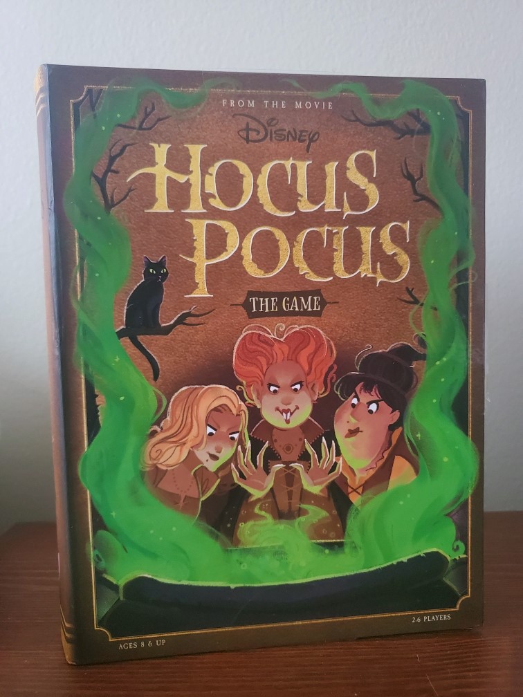 Image is a photo of Hocus Pocus The Game, board game. It shows the three Sanderson sisters on the cover. They are in front of a potion and green is coming up from the cauldron. There's a black cat sitting on a tree branch towards the left of the cover of the game. 