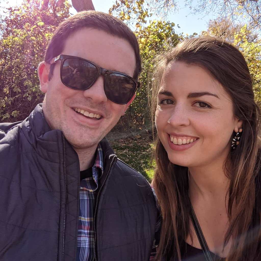 Image is a photo of me and my fiance smiling at the camera. It's a sunny day, so my husband is wearing sunglasses and a jacket. 