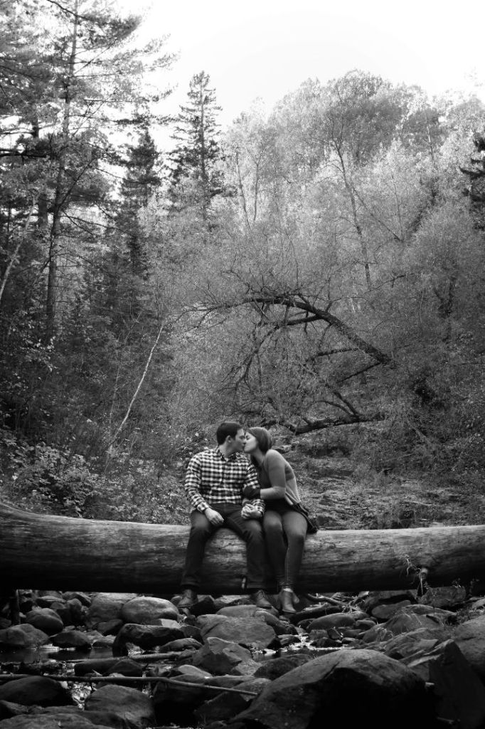 This is the black and white photo of a man and a woman kissing on a log. Trees are behind them. 