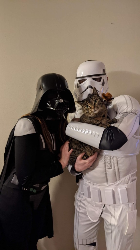Image is a photo of me, my fiance and our kitty. I'm Darth Vader, my fiance is a Storm Trooper and my fiance is holding Athena with a little harness with a cowboy mouse on it. 