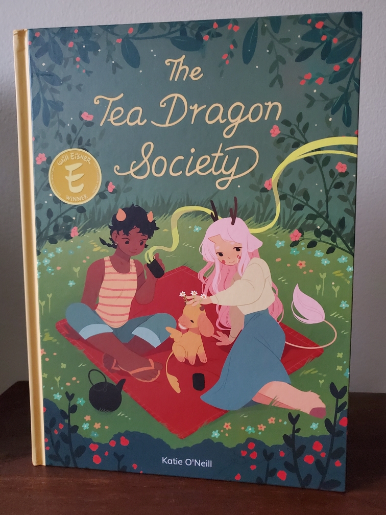Image is a graphic novel cover fro The Tea Dragon Society. It shows two cartoon like characters and they are having a picnic. On the picnic blanket is a dragon that kind of looks like a dog almost. It's orange and has a really long tail, almost like a leaf. 