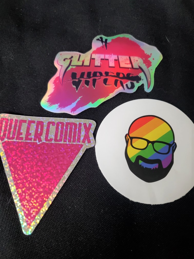 Image is a photo of three stickers. The one on the left says Queer Comix and there's a bright pink triangle pointing down. The top sticker says Glitter Vipers and the one on the right shows a bald, bearded man in rainbow. He's wearing glasses. 