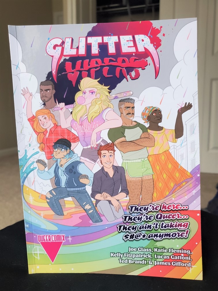 Image is a photo of a graphic novel titled Glitter Vipers. It shows seven different characters. Behind them is gray and white and in front of them everything is in color. It says" "They're here... They're Queer.... They ain't taking $#@* anymore" 