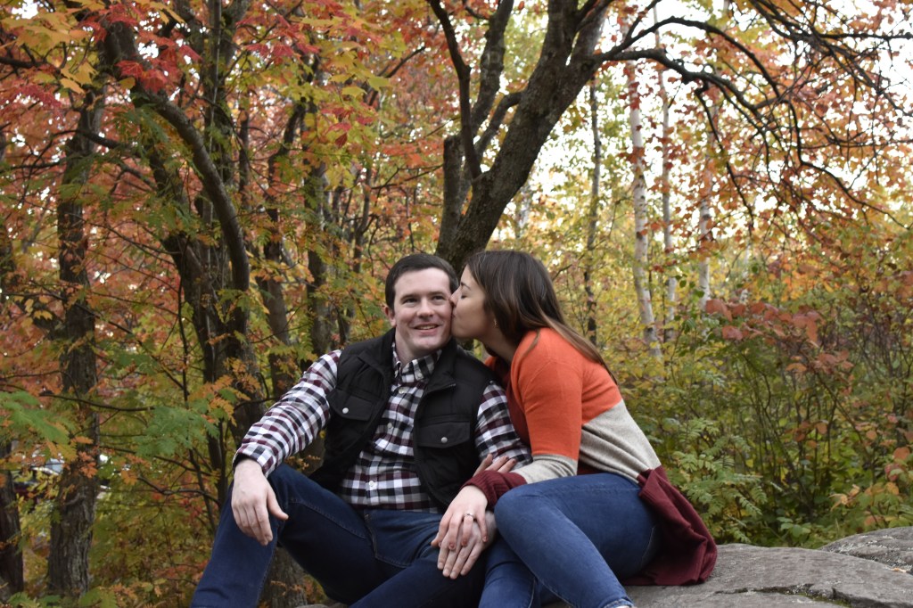 Image is a photo of me and my fiance. We are sitting on a rock and I am giving him a kiss on the cheek. Behind us, the trees are changing colors with a mixture of green, red, orange, and yellow leaves. 