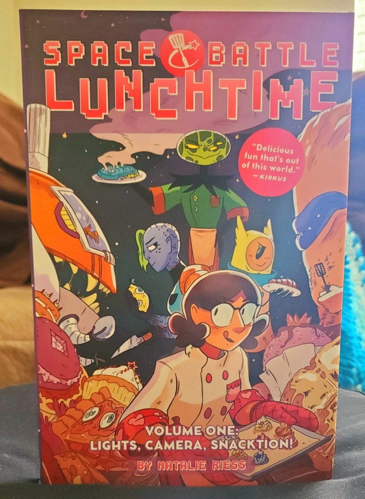 Image is a graphic novel cover for Space Battle Lunchtime. The cover has a woman on it wearing a chef uniform. She has glasses and is holding a tray of desserts. Behind her are several aliens cooking. 