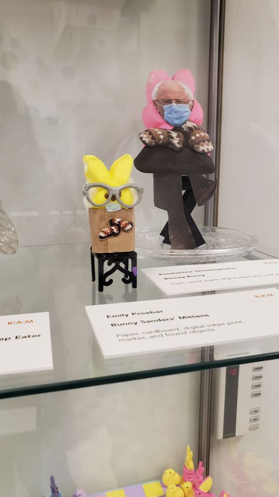 Image is a photo of an art mini with Bernie Sanders as a yellow stuffed PEEP sitting in a chair. He has silver glasses and his mittens are shown crossing over each other. 