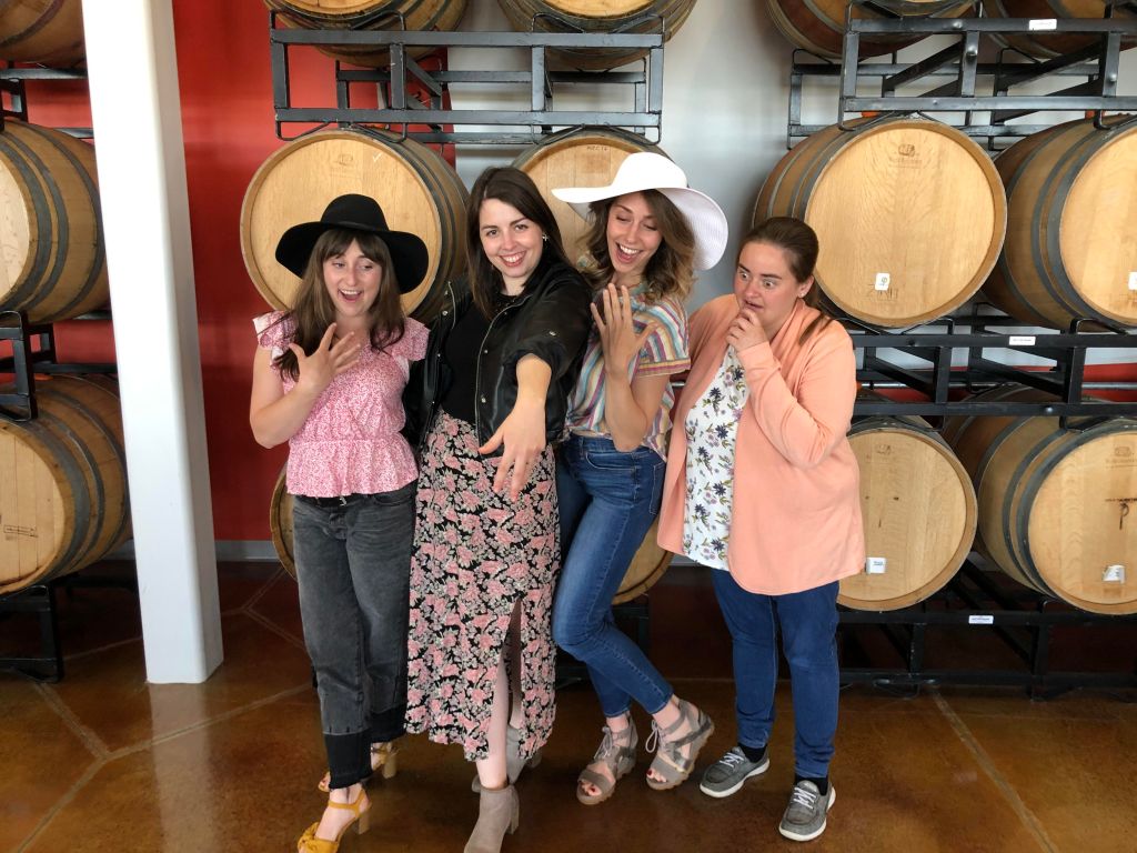 Image is a photo of me and my friends. We are in their wine room with barrels behind us. I'm showing my hand with the ring and my friends are looking at it. 