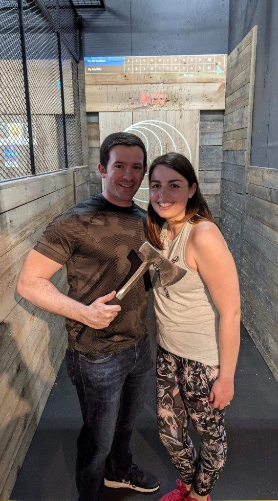 Image is a photo of me and my husband at The Burrow. Behind us is the axe throwing board. My husband is holding an axe. 