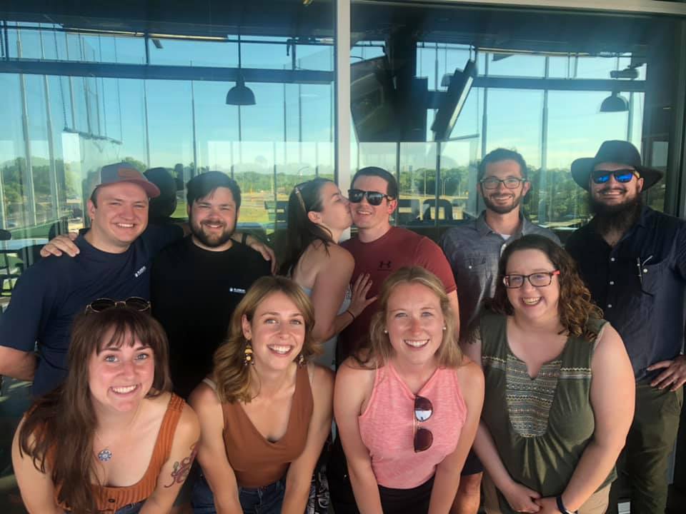 Image is a photo of 10 people, five women and five men. In the back, a woman is kissing a man on the cheek. 