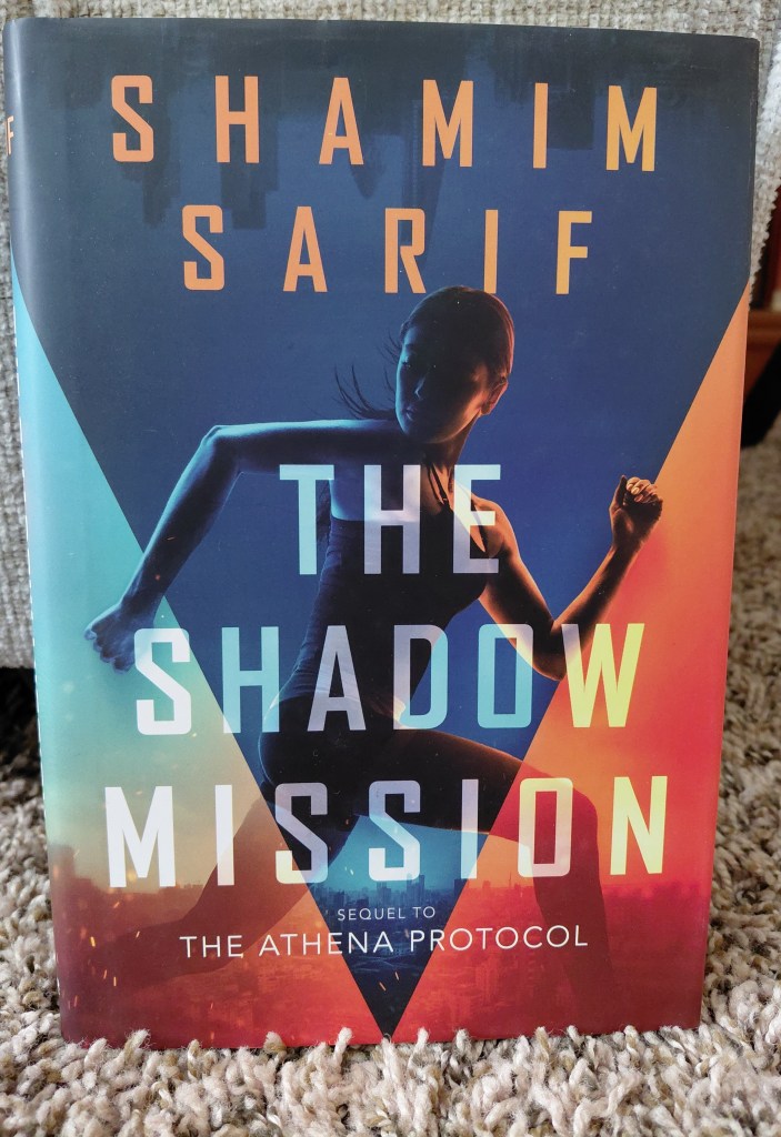 This is an image of a book cover for The Shadow Mission by Shamim Sharif. The book cover is a mix of dark blue, light blue and orange. There's a figure of a woman running, but she's looking behind her. 