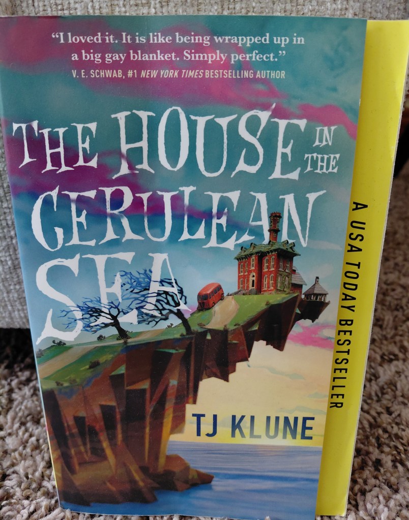 Image is a book cover for the book The House in the Cerulean Sea by TJ Klune. It shows a huge red brick house on the edge of a cliff. There's a red double decker bus parked outside. 