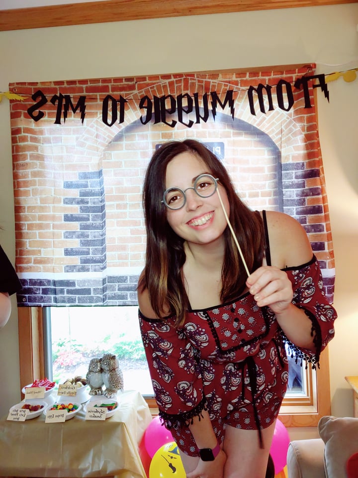 Image is a photo of me smiling at the camera. She's holding in her left hand a pair of paper glasses that are held up to her eyes. 