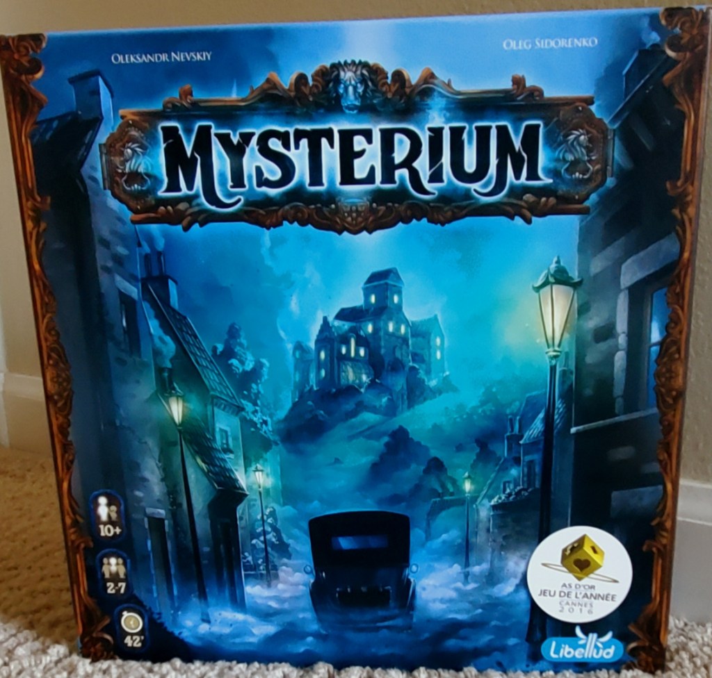 Image is a photo that I took of my copy of the board game, Mysterium. The board game shows an old car driving up to a mansion. The mansion is straight off to the distance. There appears to be fog going on the road. On both sides of the road are several buildings. 