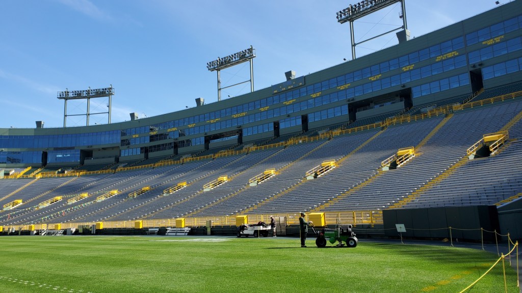 Image is a photo of Lambeau Field. This photo is taken from the field and looking up the stands. The bleechers are silver and the aisles are painted bright yellow. There's someone on the field pushing what appears to be a mini tractor or lawn mower. 
