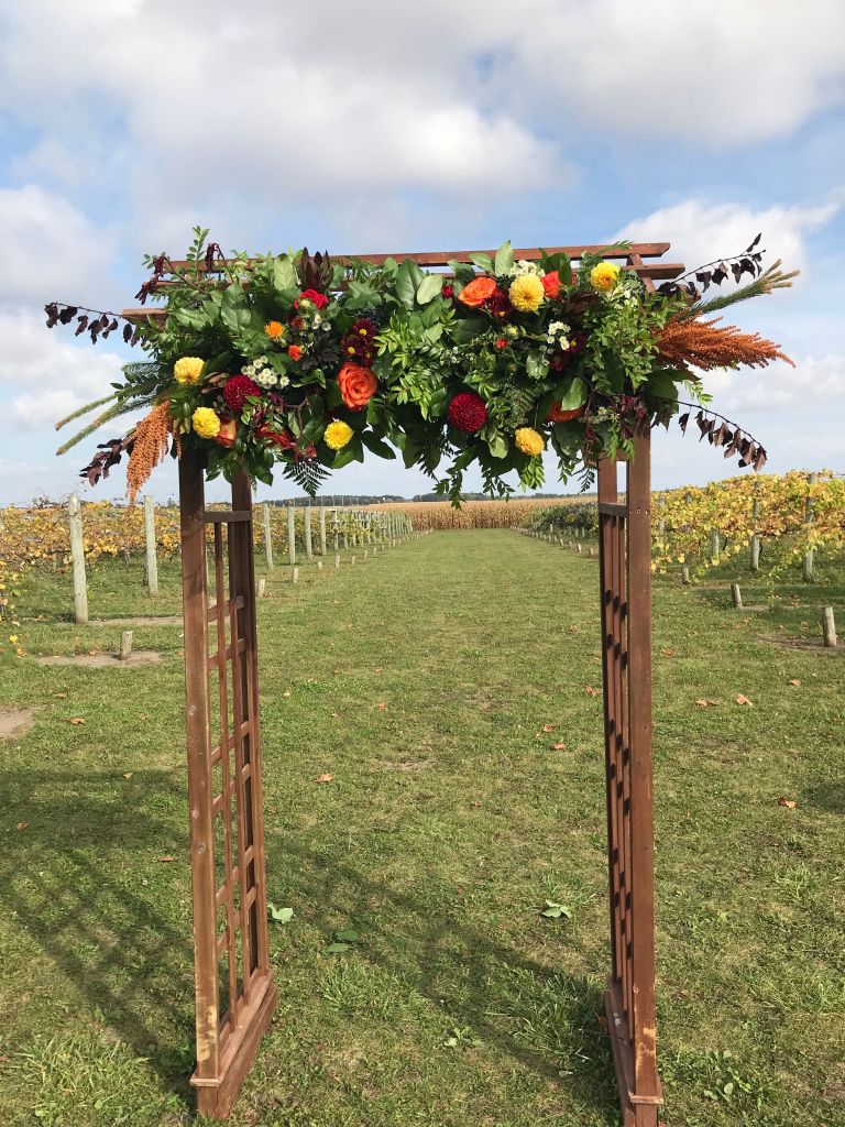 Image is a photo of a wedding arch. There's a huge floral arrangement on the front. It has roses in orange, dark blue, and bright yellow. There are leaves and ferns sticking out of it as well. On either side of the arch is the vineyard. 
