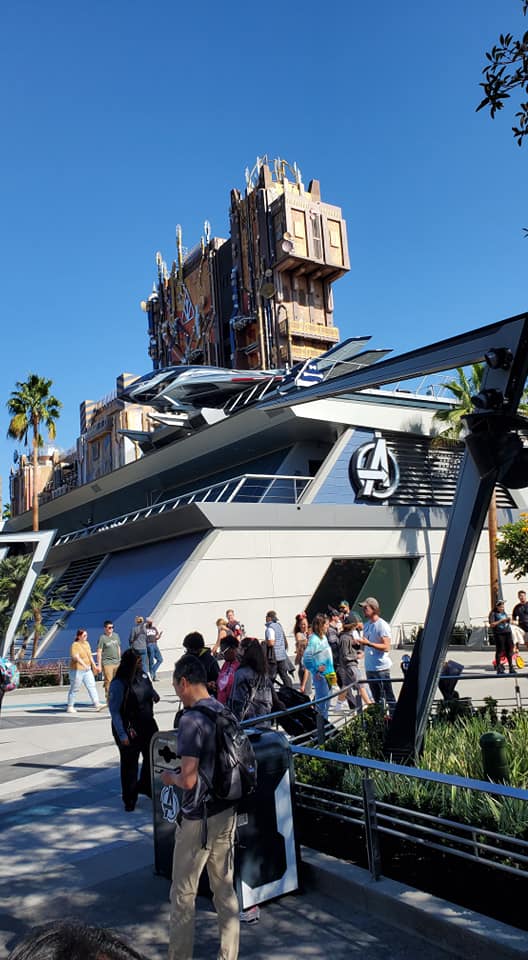 Image is a photo of Avengers Campus. There's a building with the A on it. There are people waiting outside of the building. 