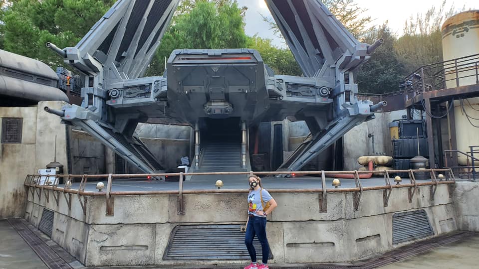Image is a photo of me posing in front of Kylo Ren's ship. There's a big ship on a pedestal and I'm standing in front of it with a hand on my hip. 
