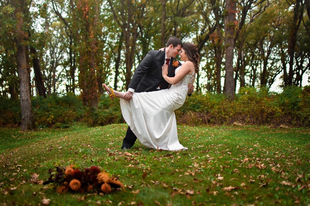 Image is a photo of me and my husband. He is lowering me into a dip with his hand on my lower back. I have my leg kicking out. My arms are wrapped around him. Behind us is a beautiful forest and leaves everywhere. 