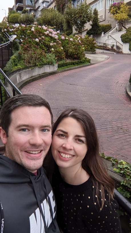 Image is a photo of me and my husband smiling at the camera. Behind us is a curvy road. 