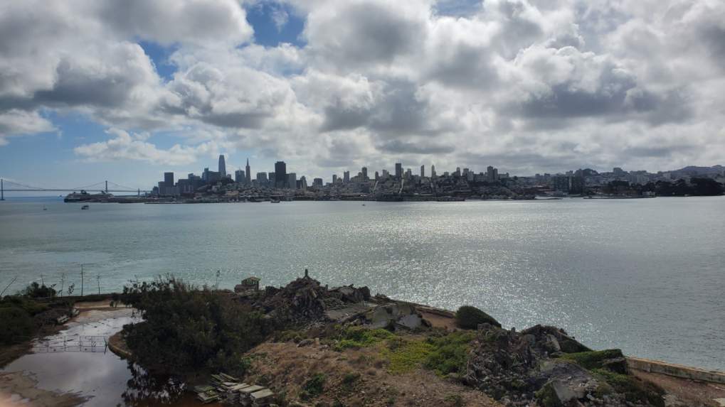Image is a photo of the San Fran skyline from Alcatrax. It shows a rock formation, a body of water, and the San Fran skyline in the distance. 
