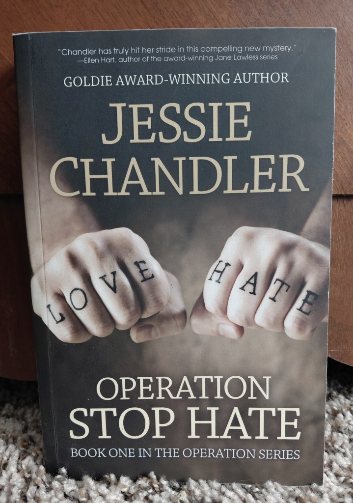 Image is a photo of a book called Operation Stop Hate by Jessie Chandler. The book cover is brown. It shows two hands with the hands clutched into fists. Written on the knuckles of one hand is "Love" in black and on the right fist knuckles is the word "Hate" in black. 